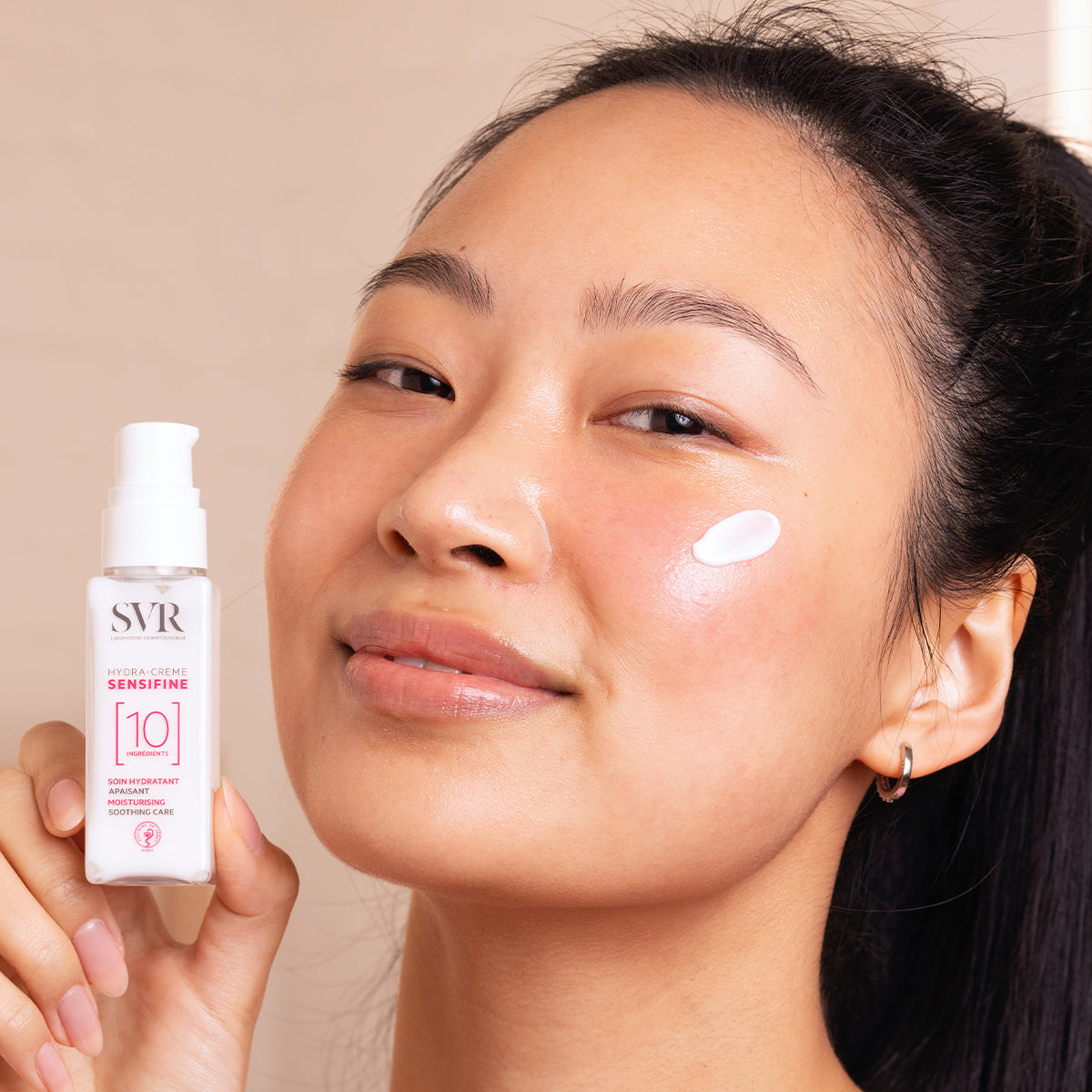 Mujer sonriente sosteniendo el envase blanco de SVR Sensifine Hydra-Creme con una gota de crema hidratante en su mejilla.