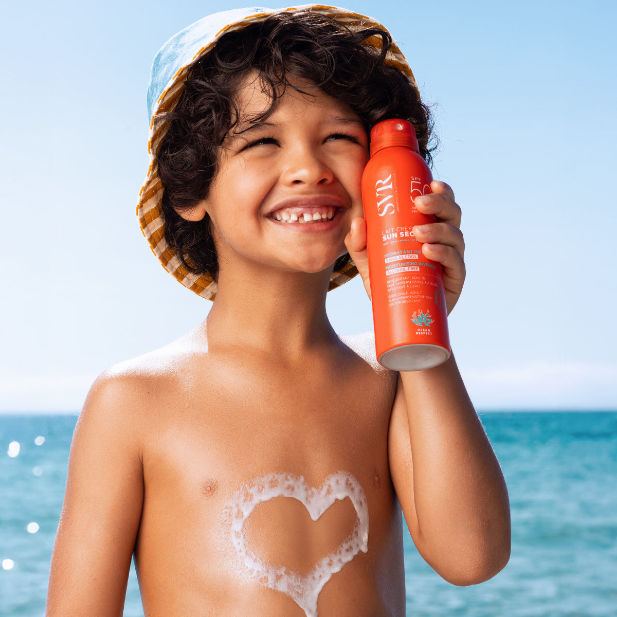 Niño sonriente con sombrero de sol sosteniendo un spray solar SVR Sun Secure SPF50+ frente al mar con un corazón de espuma.